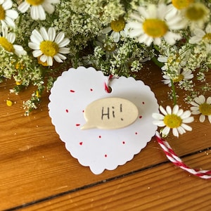 Könnte beinhalten: Ein weißes herzförmiges Etikett mit roten Punkten und einer kleinen Keramik-Sprechblase mit der Aufschrift "Hi!". Das Etikett ist mit rot-weißem Bindfaden gebunden und von Gänseblümchen und anderen weißen Blumen umgeben.