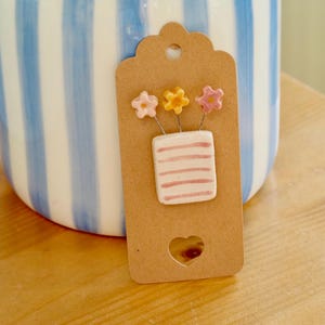 Op de afbeelding: Een decoratief label met een hartvormige uitsparing toont een keramische vaas met drie kleine bloemen. De vaas is wit met roze strepen en de bloemen zijn roze en geel. Op de achtergrond staat een blauw-wit gestreepte keramische vaas.