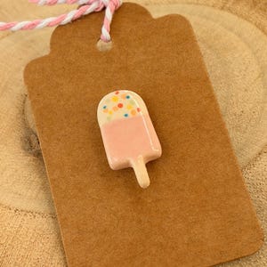 Könnte beinhalten: Eine rosa Eis am Stiel-förmige Anstecknadel mit einem weißen Oberteil, das mit bunten Streuseln bedeckt ist. Die Anstecknadel befindet sich auf einem braunen Anhänger mit einer rosa-weißen Schnur. Ein lustiges Accessoire.