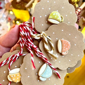 Könnte beinhalten: Eine Sammlung handgefertigter Geschenkanhänger mit gewelltem Rand und Polka-Dot-Muster. Jeder Anhänger hat eine Miniatur-Keramik-Teetasse. Die Teetassen sind in verschiedenen Farben, darunter Gelb, Orange und Blau, und sind mit rot-weißem Garn zusammengebunden.
