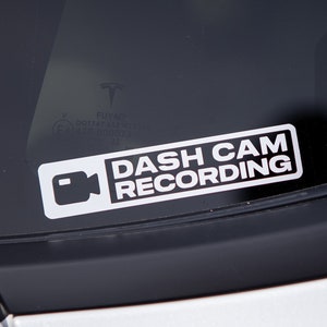 Può includere: Un adesivo bianco con testo nero che dice "DASH CAM RECORDING" e un'icona di una telecamera nera.