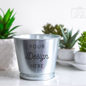May include: A silver metal planter with the text "YOUR Design HERE" on the side. The planter is sitting on a white surface with three small white planters with green plants.