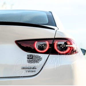 Peut inclure: Voiture blanche avec un autocollant de drapeau américain noir et blanc qui dit "FDT" à l'arrière. La voiture a également un emblème "Skyactiv-G Turbo".