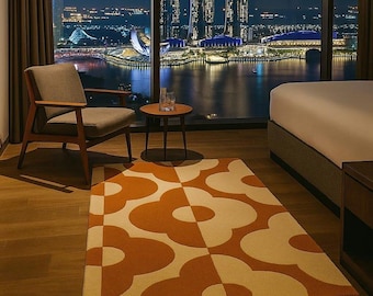 May include: A rectangular rug with a floral pattern in shades of orange and cream. The rug is placed on a wooden floor in a room with a view of a city skyline at night. A wooden chair and side table are also in the room.