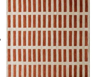 May include: Rectangular rug with a repeating pattern of vertical, rust-colored rectangles against a cream background. The rug's design is symmetrical, creating a grid-like appearance. The rug is suitable for a living room or bedroom.