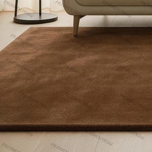 May include: A rectangular, solid brown rug with a soft, plush texture. The rug is placed on a light-coloured wooden floor, with a portion of a beige sofa and a black side table visible in the background. The rug appears to be new.