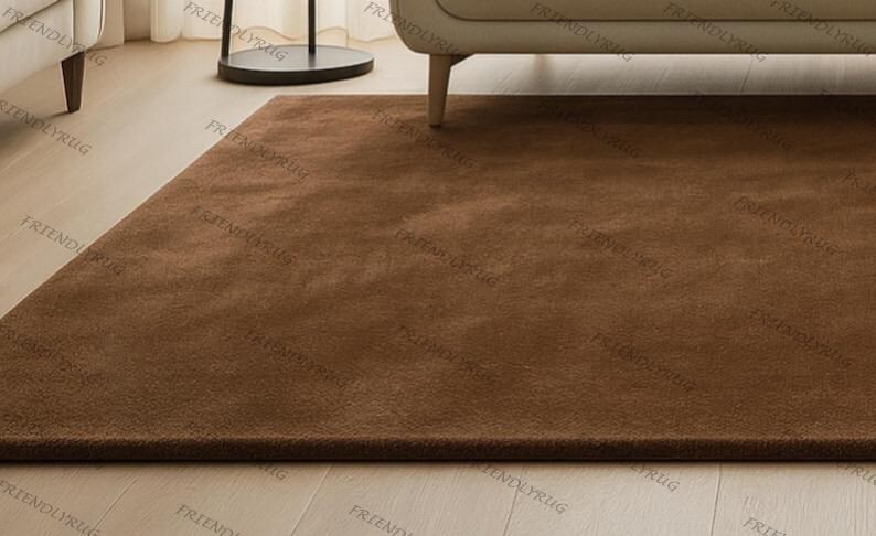 May include: A rectangular, solid brown rug with a soft, plush texture. The rug is placed on a light-coloured wooden floor, with a portion of a beige sofa and a black side table visible in the background. The rug appears to be new.