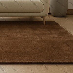 May include: A rectangular, brown rug with a soft, plush texture. The rug is placed on a light-coloured wooden floor, with a portion of a beige sofa visible in the background. The rug's colour is a warm, earthy tone.