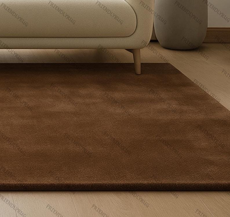 May include: A rectangular, brown rug with a soft, plush texture. The rug is placed on a light-coloured wooden floor, with a portion of a beige sofa visible in the background. The rug's colour is a warm, earthy tone.