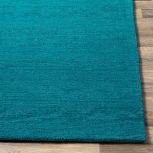 May include: A close-up of a teal rug with a textured surface. The rug is rectangular and has a solid colour. The rug is placed on a wooden floor.