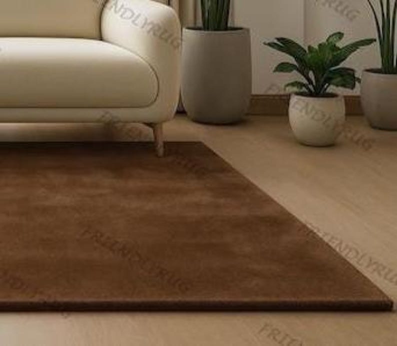 May include: A rectangular brown rug on a light wood floor, in front of a cream sofa. Potted plants are in the background. The rug's colour is a rich, earthy tone.
