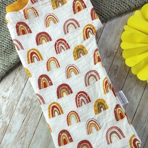 May include: A white muslin baby swaddle with a rainbow pattern in shades of red, orange, yellow, and green. The swaddle has a yellow trim and a small tag that says "Handmade with love".