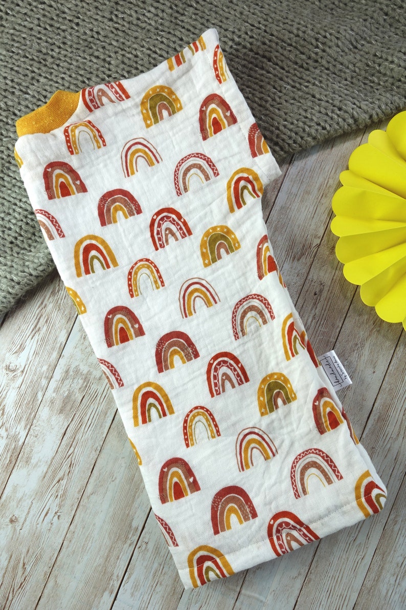 May include: A white muslin baby swaddle with a rainbow pattern in shades of red, orange, yellow, and green. The swaddle has a yellow trim and a small tag that says "Handmade with love".