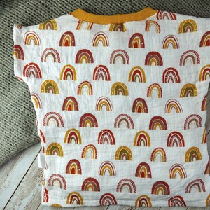 May include: White baby t-shirt with a colourful rainbow pattern. The shirt has a yellow collar and short sleeves.