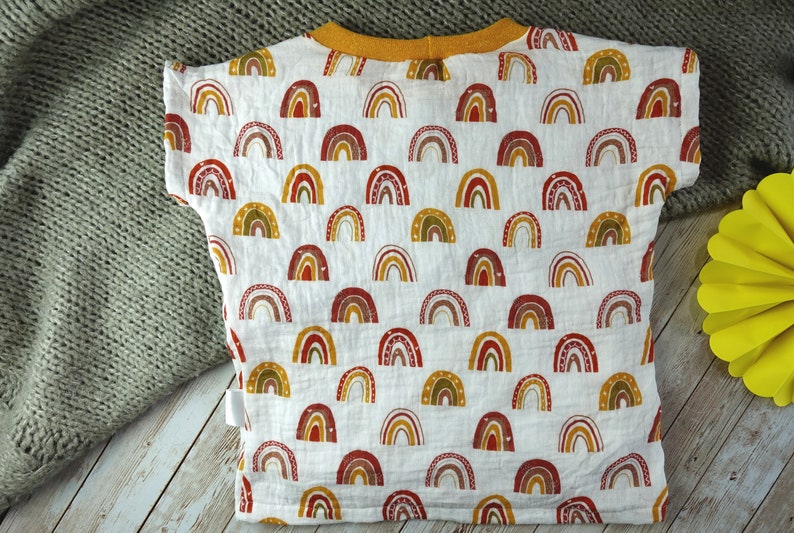May include: White baby t-shirt with a colourful rainbow pattern. The shirt has a yellow collar and short sleeves.