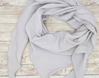 May include: A light grey cotton gauze scarf with a textured weave. The scarf is folded in a triangular shape and is lying on a light wood surface.