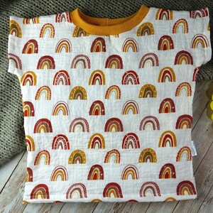 May include: A white t-shirt with a pattern of colourful rainbows. The shirt has a yellow collar and short sleeves.