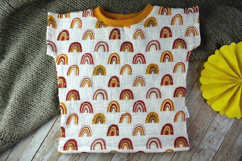 May include: A white t-shirt with a pattern of colourful rainbows. The shirt has a yellow collar and short sleeves.