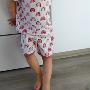 May include: A child wearing a white and pink short-sleeved shirt and matching shorts with a rainbow pattern. The shirt and shorts have a pink trim.