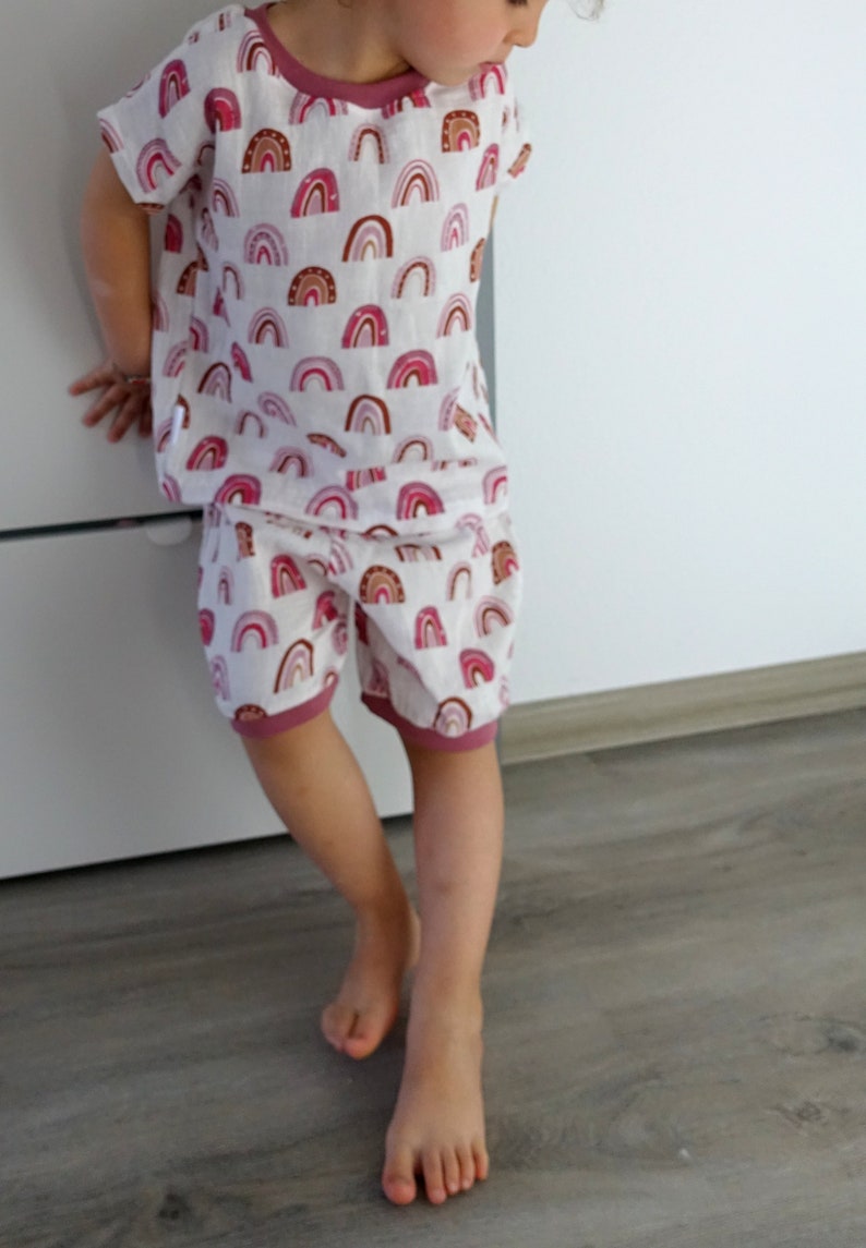May include: A child wearing a white and pink short-sleeved shirt and matching shorts with a rainbow pattern. The shirt and shorts have a pink trim.