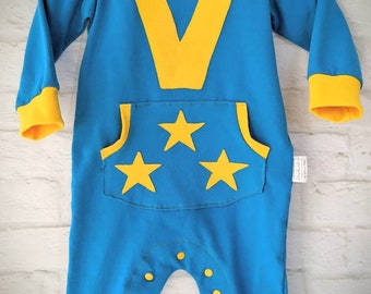May include: A blue baby onesie with yellow accents. The onesie has a large yellow letter V on the front and a pocket with three yellow stars. The onesie has yellow cuffs and a yellow trim around the neckline.