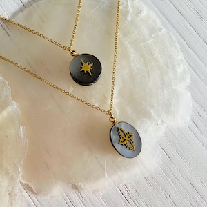 May include: Two gold necklaces with pendants. The top necklace has a round black pendant with a gold star. The bottom necklace has a round black pendant with a gold floral design.