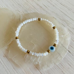 Peut inclure: Bracelet en perles de coquillage blanc avec un charme œil maléfique doré. Le bracelet est sur un fond blanc.