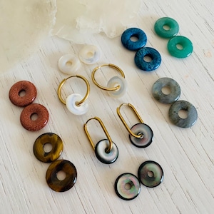 Pode incluir: Conjunto de brincos de argola de pedras preciosas e conchas. Os brincos são feitos com vários materiais, incluindo ouro, prata e pedras naturais. Os brincos são exibidos em uma superfície de madeira branca.