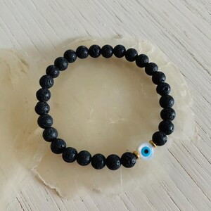 Peut inclure: Bracelet en perles de roche volcanique noire avec un charme œil maléfique blanc. Le charme a un œil bleu au centre.