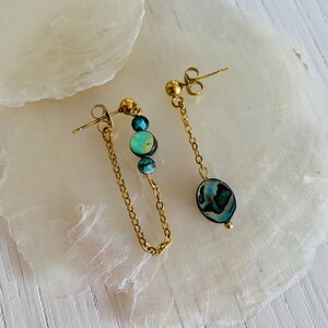 May include: A pair of gold-colored earrings. One earring features a chain with three turquoise and blue beads. The other earring has a single oval-shaped abalone shell bead. Both earrings have a gold-colored stud.
