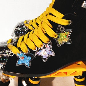 May include: Black roller skates with yellow laces and clear star-shaped charms with colorful glitter. The skates have yellow wheels and a black frame.