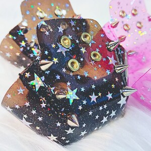 以下が含まれることがあります： ホログラフィックな星柄と金属製のスパイクが特徴のリストカフ3点。ブラック、ブラウン、ピンクの半透明素材で作られており、それぞれ2つのゴールドトーンの金属製アイレットでサイズ調整が可能です。