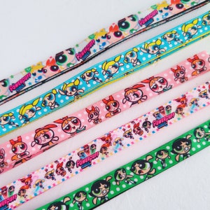 Peut inclure: Lanières Powerpuff Girls de différentes couleurs. Chaque lanière présente un motif répété des personnages des Powerpuff Girls et le texte "Powerpuff Girls". Les couleurs incluent le bleu clair, le rose, le vert et le blanc.