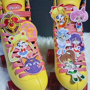 May include: Pair of yellow Moxi roller skates with pink laces and red wheels. The skates are decorated with colorful embroidered patches of anime characters. The Moxi logo is visible on the tongue of the skates.