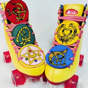 May include: Pair of yellow Moxi roller skates with pink laces and red wheels. Each skate features multiple round patches with colorful embroidered designs. The patches are red, green, pink, yellow, and blue.