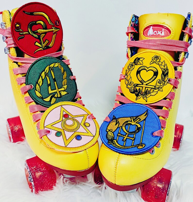 May include: Pair of yellow Moxi roller skates with pink laces and red wheels. Each skate features multiple round patches with colorful embroidered designs. The patches are red, green, pink, yellow, and blue.
