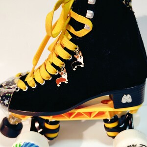 May include: Black roller skates with yellow laces and a leopard print trim. The skates have a black and white patterned wheel with the text "Park 74" on it. The skates have two small charms attached to the laces, each featuring a cartoon corgi.
