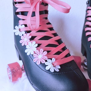 May include: Black roller skates with pink laces and wheels. The skates are decorated with small, white and pink flower appliqu&eacute;s. The word "IMPA" is visible on the side of the skate.
