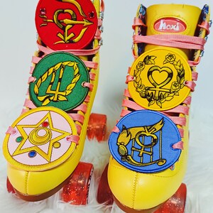 May include: A pair of yellow roller skates with pink laces and decorative patches. The patches are round with various symbols and colors, including red, green, pink, yellow, and blue. The wheels are red and the skates have the word "hoxi" on the tongue.