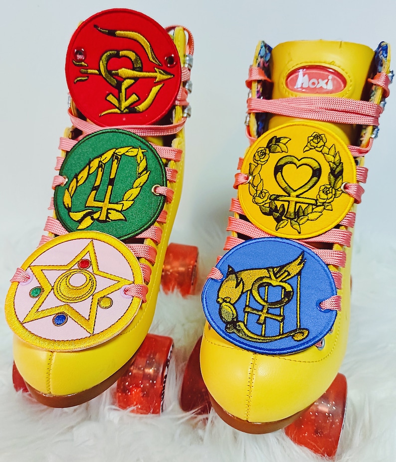 May include: A pair of yellow roller skates with pink laces and decorative patches. The patches are round with various symbols and colors, including red, green, pink, yellow, and blue. The wheels are red and the skates have the word "hoxi" on the tongue.