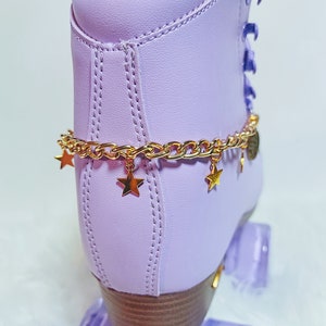 May include: Purple roller skates with gold chain and star charms. The skates have a light purple boot with a brown wooden base and clear purple wheels.