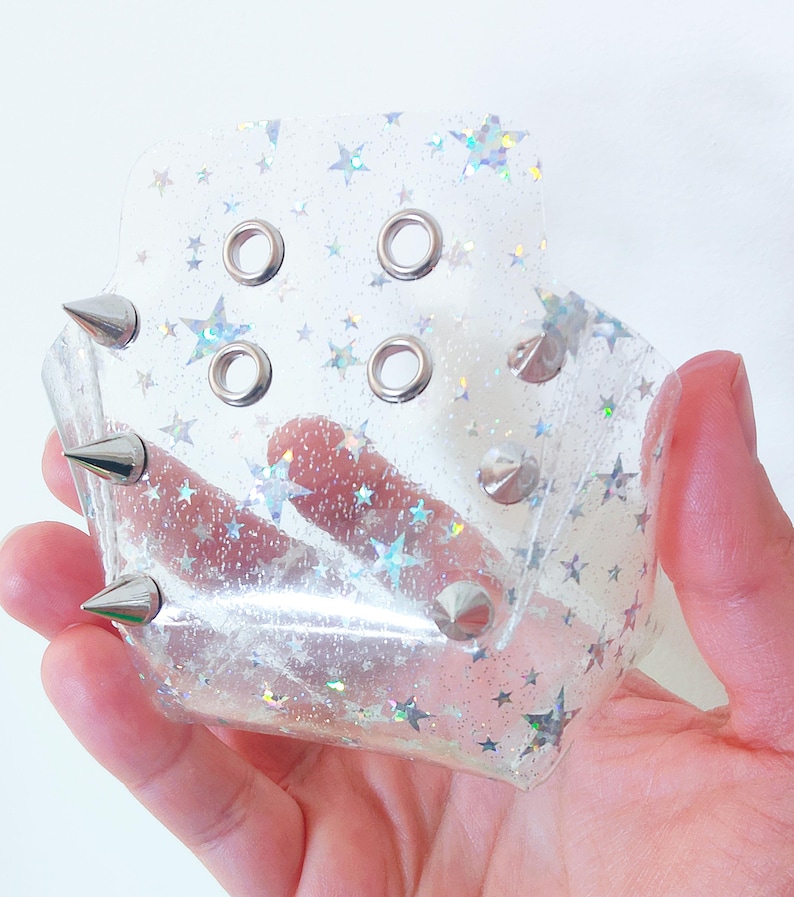 以下が含まれることがあります： ホログラフィックな星柄と縁に沿った銀色の金属製スパイクが特徴の透明で柔軟なカフ。アクセサリーの前面には、調整用の四角い配置で4つの円形の金属製アイレットが施されています。