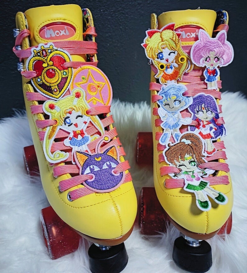 May include: Pair of yellow Moxi roller skates with pink laces and red wheels. The skates are decorated with colorful embroidered patches of anime characters. The Moxi logo is visible on the tongue of the skates.