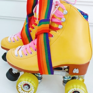 May include: Yellow roller skates with pink laces and a rainbow strap. The wheels are yellow with white lettering that says "EnerG" on them.