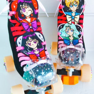 May include: Pair of black roller skates with red laces and pink bows. The skates feature embroidered anime character designs and clear toe stops with a star pattern. The wheels are beige and the stoppers are orange.
