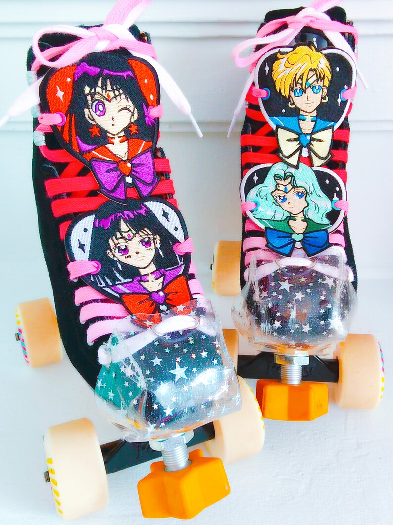 May include: Pair of black roller skates with red laces and pink bows. The skates feature embroidered anime character designs and clear toe stops with a star pattern. The wheels are beige and the stoppers are orange.