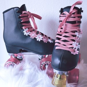 May include: Black roller skates with pink laces and daisy accents. The skates have pink wheels and are set against a white, fluffy background. The laces are tied in bows, and the wheels have glitter.