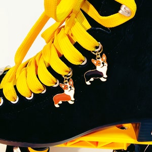 May include: Black roller skates with yellow laces and two small dog charms attached to the laces. The charms are of a black and white corgi and a brown and white corgi.