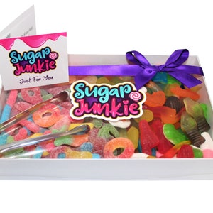 May include: A white gift box filled with colorful gummy candies. The box is tied with a purple ribbon and has a "Sugar Junkie" logo on the front. The box also includes a pair of silver tongs for picking up the candies.