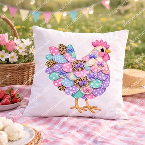 May include: White square cushion with a colourful chicken design. The chicken has a pink comb, yellow beak, and a body made of various patterned fabrics, including polka dots, leopard print, and gingham. Purple bows accent the design. The cushion is on a pink and white checkered picnic blanket.